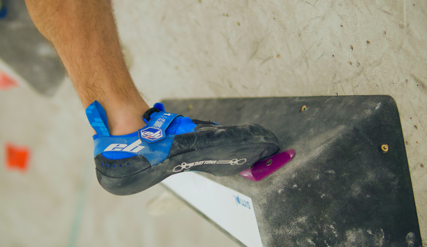EB Nebula Climbing shoes in blue and black. The climber is on an indoor climbing wall pushing off on his right foot on a small purple foot-hold.