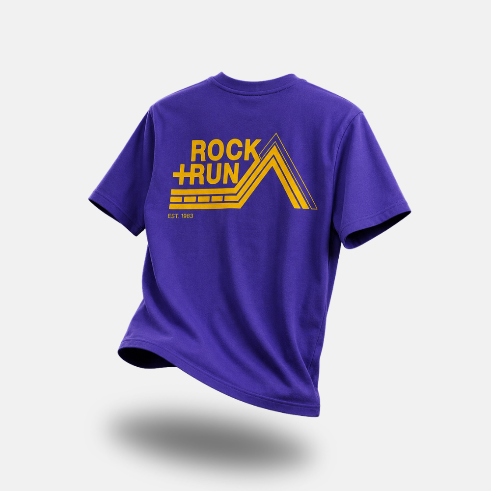 A purple short-sleeve t-shirt with a yellow graphic on the back reading 'ROCK + RUN' and 'EST. 1983', shown floating against a light gray background.