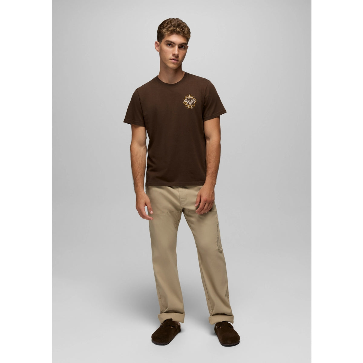 Man wearing a brown t-shirt with a logo and beige pants on a gray background